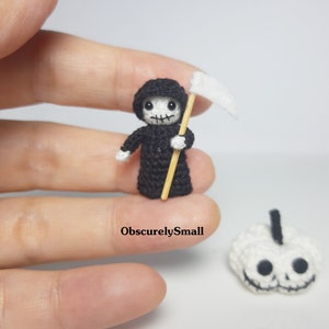 May include: A small crocheted Grim Reaper figure holding a scythe, with a white crocheted pumpkin in the background. The figure is wearing a black robe and has a white face with black eyes and a black mouth.