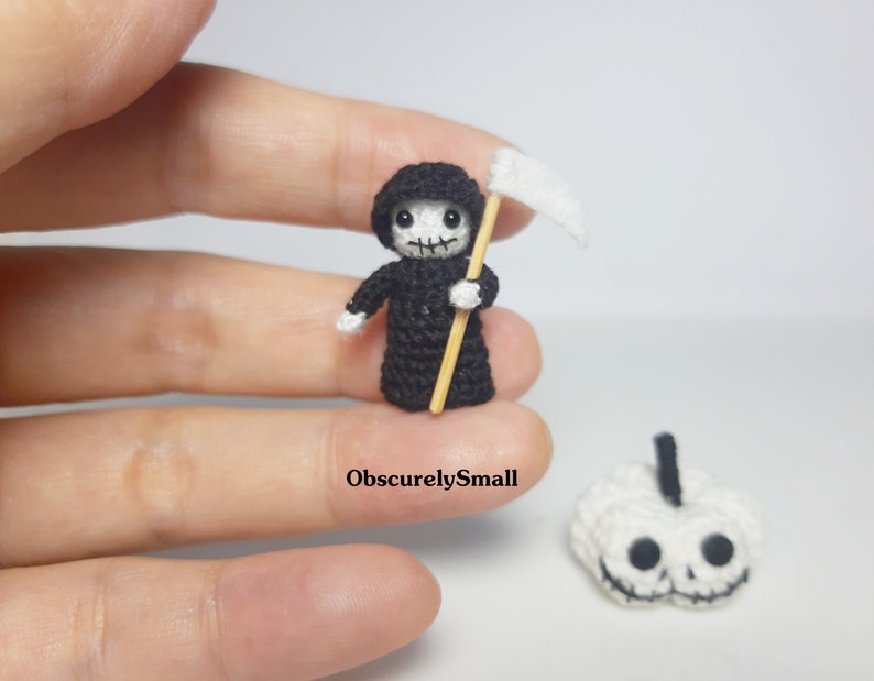 May include: A small crocheted Grim Reaper figure holding a scythe, with a white crocheted pumpkin in the background. The figure is wearing a black robe and has a white face with black eyes and a black mouth.