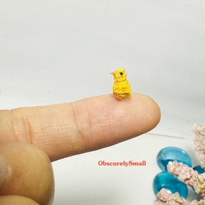 May include: A tiny yellow crocheted chick with orange beak and legs, perched on a finger. The chick is very small, almost the size of a fingernail.