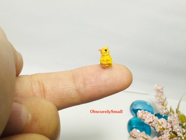 May include: A tiny yellow crocheted chick with orange beak and legs, perched on a finger. The chick is very small, almost the size of a fingernail.
