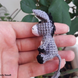 Puede incluir: Un peque&ntilde;o burro de crochet gris con ojos negros y cola negra. El burro est&aacute; sentado en la mano de una persona.