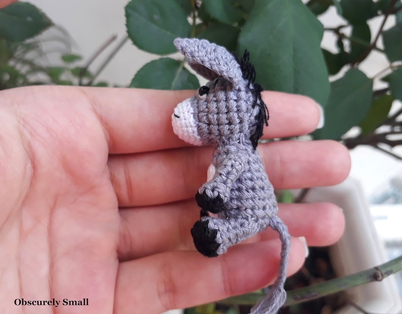 Puede incluir: Un peque&ntilde;o burro de crochet gris con ojos negros y cola negra. El burro est&aacute; sentado en la mano de una persona.