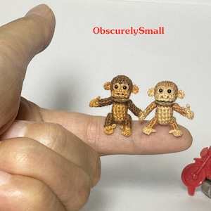 May include: Two miniature crochet monkeys, one brown and one tan, are displayed on a finger. The monkeys are holding hands and have detailed facial features. A red toy motorcycle is in the background. The text "ObscurelySmall" is in the upper right corner.