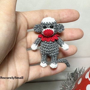 Puede incluir: Un peque&ntilde;o mono de crochet gris con la nariz roja y un collar de crochet blanco y rojo. El mono est&aacute; sentado en una mano.