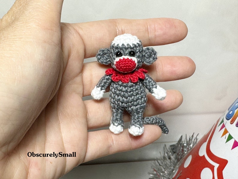 Puede incluir: Un peque&ntilde;o mono de crochet gris con la nariz roja y un collar de crochet blanco y rojo. El mono est&aacute; sentado en una mano.