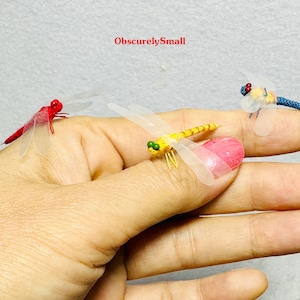 May include: Three miniature dragonfly figurines, one red, one yellow, and one blue, are displayed on a hand. The dragonflies are made of fabric and have delicate wings.