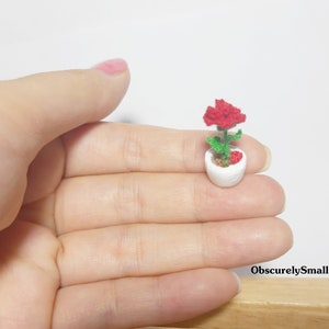 May include: A miniature white ceramic flower pot with a red crocheted flower and green leaves. The pot is held in a hand, showcasing the small size of the handcrafted item. The text "ObscurelySmall" is visible.