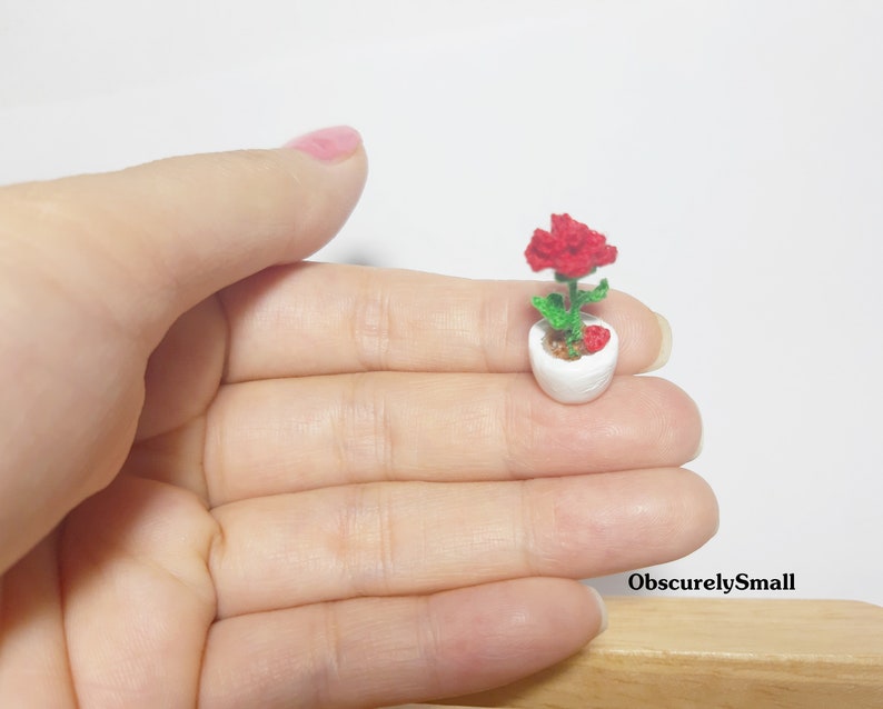 May include: A miniature white ceramic flower pot with a red crocheted flower and green leaves. The pot is held in a hand, showcasing the small size of the handcrafted item. The text "ObscurelySmall" is visible.