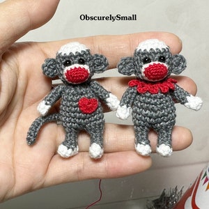 Puede incluir: Dos peque&ntilde;os monos de crochet grises en forma de calcet&iacute;n con narices rojas y corazones rojos en el pecho. Un mono tiene un collar rojo de crochet.