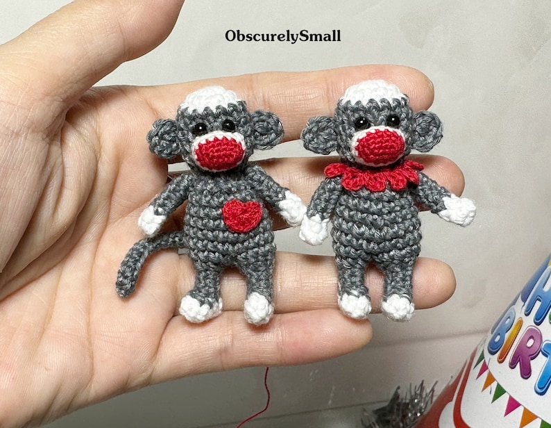 Puede incluir: Dos peque&ntilde;os monos de crochet grises en forma de calcet&iacute;n con narices rojas y corazones rojos en el pecho. Un mono tiene un collar rojo de crochet.