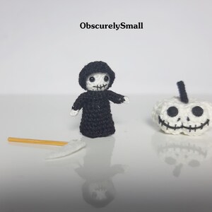 May include: Crocheted Halloween-themed figures, including a small Grim Reaper in a black robe and a white pumpkin with a stitched face. A small scythe lies nearby. The text "ObscurelySmall" is visible.