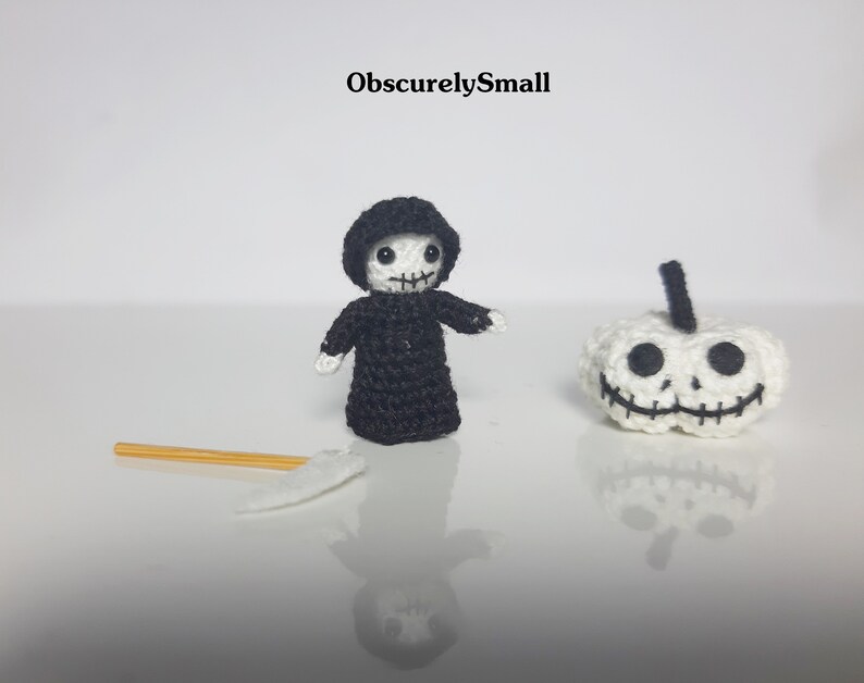 May include: Crocheted Halloween-themed figures, including a small Grim Reaper in a black robe and a white pumpkin with a stitched face. A small scythe lies nearby. The text "ObscurelySmall" is visible.