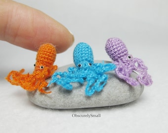 May include: Three crocheted octopus figurines, one orange, one blue, and one purple, are sitting on a gray stone. The octopus figurines are very small, and the stone is about the size of a fingertip.