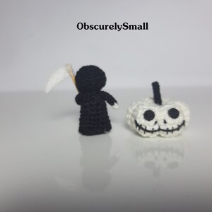 May include: A black crocheted figure of the Grim Reaper holding a white scythe, standing next to a white crocheted pumpkin with a black stitched-on face.