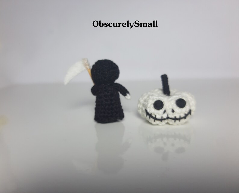 May include: A black crocheted figure of the Grim Reaper holding a white scythe, standing next to a white crocheted pumpkin with a black stitched-on face.
