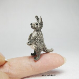 May include: A small gray crocheted kangaroo with a joey in its pouch. The kangaroo is standing on a finger.
