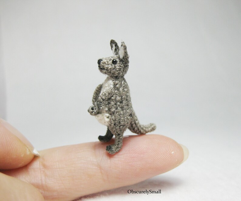 May include: A small gray crocheted kangaroo with a joey in its pouch. The kangaroo is standing on a finger.