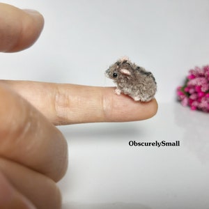 May include: A tiny gray and brown crocheted mouse with pink ears sits on a finger. The mouse is very small, about the size of a fingernail.