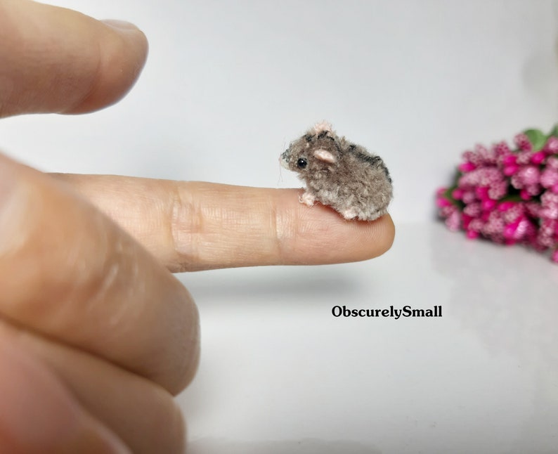 May include: A tiny gray and brown crocheted mouse with pink ears sits on a finger. The mouse is very small, about the size of a fingernail.