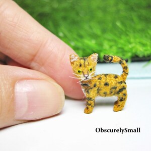 Pu&ograve; includere: Un piccolo gatto tabby giallo e nero all'uncinetto con occhi verdi. Il gatto &egrave; in piedi su una superficie bianca.