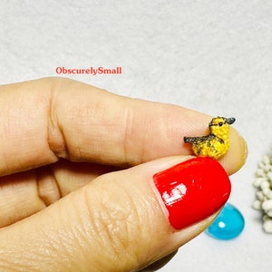 Op de afbeelding: Een klein gehaakt geel eendje met een zwart hoofd en snavel. Het eendje wordt vastgehouden door een hand met rode nagellak.