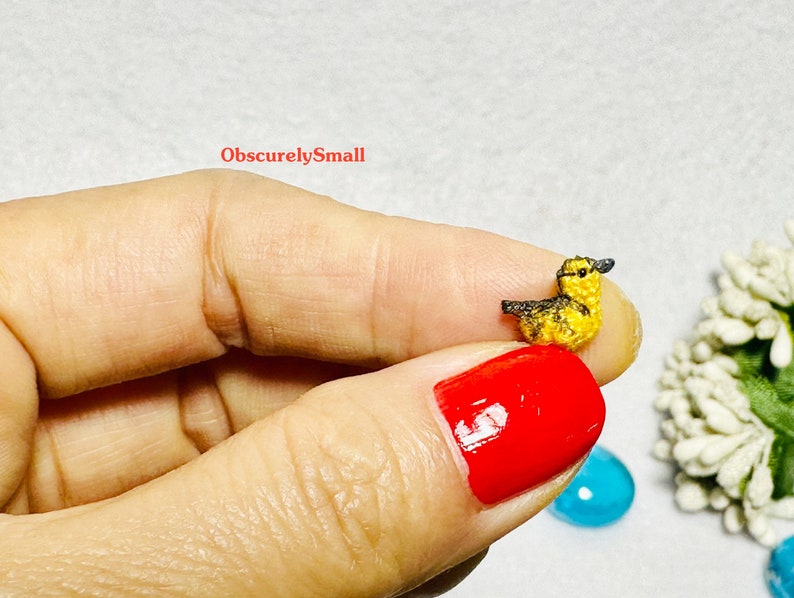 Op de afbeelding: Een klein gehaakt geel eendje met een zwart hoofd en snavel. Het eendje wordt vastgehouden door een hand met rode nagellak.