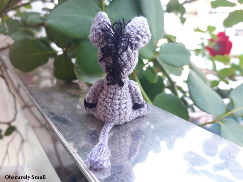 Puede incluir: Un peque&ntilde;o burro de crochet gris con hilo negro para la crin y la cola. El burro est&aacute; sentado en una superficie plateada.