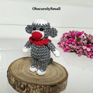 Puede incluir: Un peque&ntilde;o mono de crochet gris con un collar de crochet rojo y blanco. El mono est&aacute; de pie sobre una base de madera.