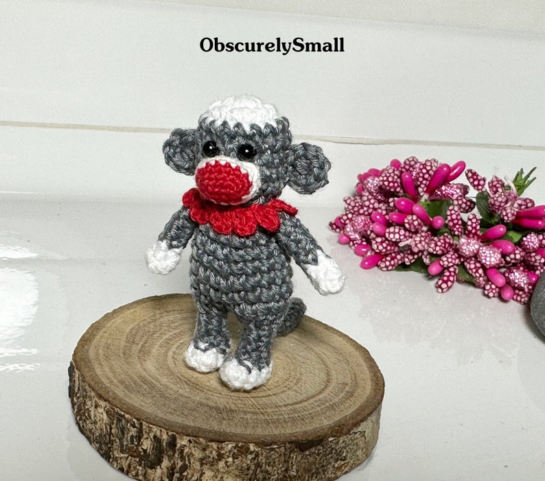 Puede incluir: Un peque&ntilde;o mono de crochet gris con un collar de crochet rojo y blanco. El mono est&aacute; de pie sobre una base de madera.