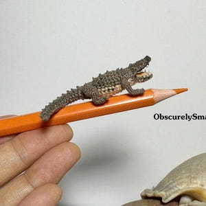 May include: A small crocheted alligator with an open mouth, perched on a yellow pencil. The alligator is brown and gray with a white belly. The pencil is held by a hand, and the text "ObscurelySmall" is visible in the background.