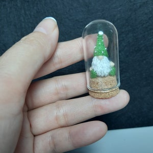 May include: A small green gnome wearing a green hat sits inside a clear glass jar with a cork lid. The gnome is sitting on a small piece of cork.