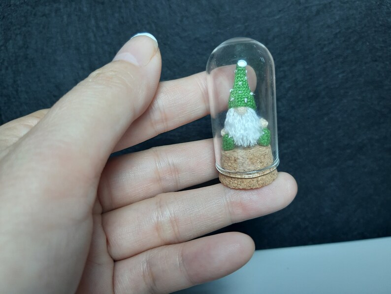 May include: A small green gnome wearing a green hat sits inside a clear glass jar with a cork lid. The gnome is sitting on a small piece of cork.