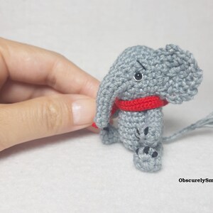 May include: A small gray crocheted elephant figurine featuring a bright red scarf around its neck, black bead eyes, and black stitched details on its feet. The text ObscurelySmall is printed in the bottom right corner of the image.