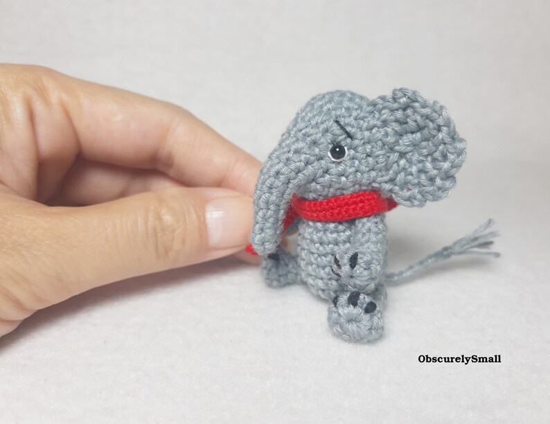 May include: A small gray crocheted elephant figurine featuring a bright red scarf around its neck, black bead eyes, and black stitched details on its feet. The text ObscurelySmall is printed in the bottom right corner of the image.