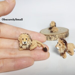 Pu&ograve; includere: Tre figurine di leone all'uncinetto. Due sono di colore beige con criniera marrone e una &egrave; beige con criniera marrone e viso bianco. I leoni sono seduti su una superficie di legno e una mano tiene uno dei leoni.