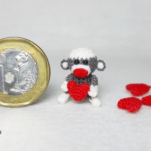 Może przedstawiać: Miniaturowa, ręcznie robiona małpka z skarpet trzymająca czerwone serce, obok monety. Małpka jest szara, biała i czerwona. Kilka małych czerwonych przedmiotów w kształcie serca jest rozrzuconych w pobliżu. Widoczny jest tekst "ObscurelySmall".