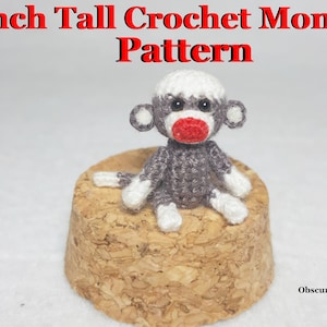 May include: A crocheted monkey toy, 1 inch tall, sitting on a cork surface. The monkey is gray with white accents and a red nose. The toy is part of a crochet pattern.