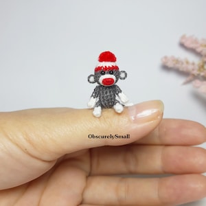 May include: A small crocheted monkey wearing a red and white hat. The monkey is gray with white accents and is sitting on a finger.