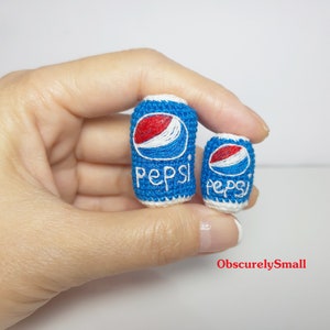Puede incluir: Dos latas de Pepsi de crochet, una más grande que la otra, se sostienen en una mano. Las latas son azules y blancas con el logotipo de Pepsi bordado.