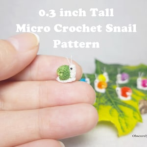 Puede incluir: Un pequeño caracol de crochet, que mide 0,8 cm de alto, se sostiene en una mano. El caracol es verde y blanco con un ojo negro. Varios otros caracoles de crochet en varios colores están sobre una hoja verde en el fondo. El texto "0.3 inch Tall Micro Crochet Snail Pattern" está en la parte superior de la imagen.