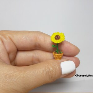 May include: A miniature crocheted sunflower in a small woven pot. The sunflower has bright yellow petals and a brown center. The pot is a warm orange color, and the stem and leaves are green. The text "ObscurelySmall" is in the bottom right corner.