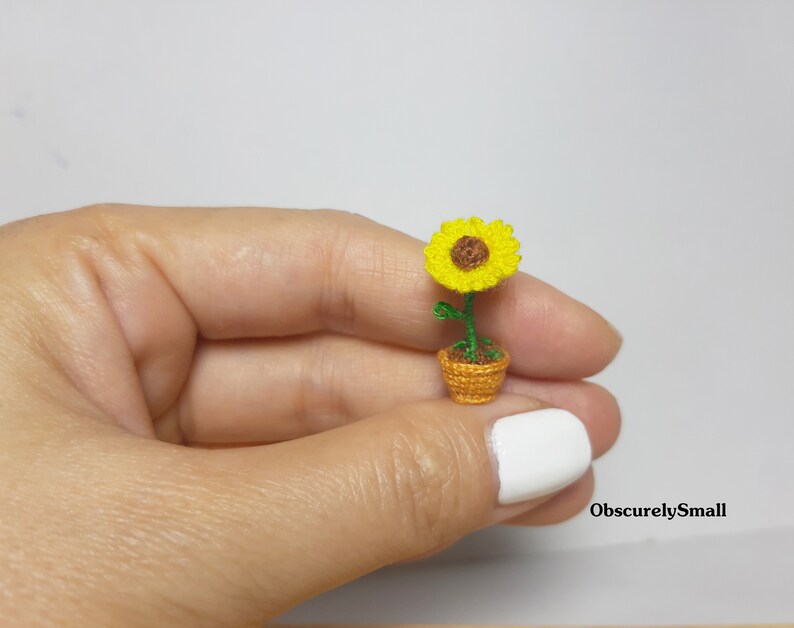 May include: A miniature crocheted sunflower in a small woven pot. The sunflower has bright yellow petals and a brown center. The pot is a warm orange color, and the stem and leaves are green. The text "ObscurelySmall" is in the bottom right corner.