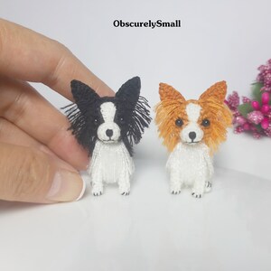 May include: Two miniature crocheted papillon dogs, one black and white and one orange and white, sitting on a white surface.