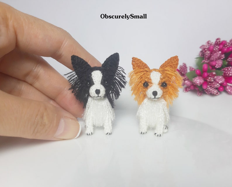 May include: Two miniature crocheted papillon dogs, one black and white and one orange and white, sitting on a white surface.