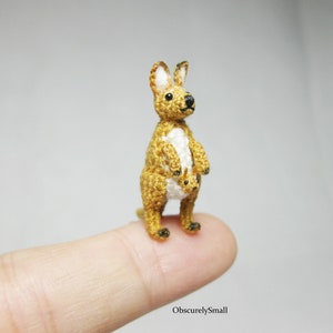May include: A small crocheted kangaroo with a joey in its pouch. The kangaroo is brown and white, and the joey is white. The kangaroo is standing on a finger.