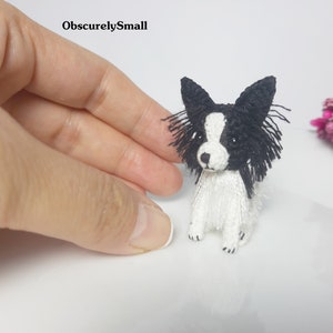 May include: A small crocheted black and white papillon dog with black eyes and a white body. The dog is sitting on a white surface.