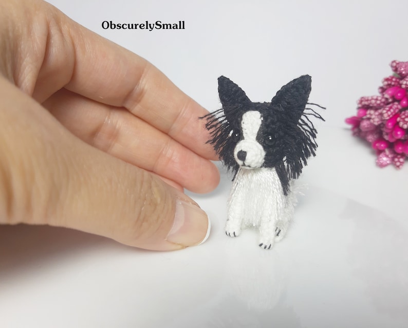 May include: A small crocheted black and white papillon dog with black eyes and a white body. The dog is sitting on a white surface.