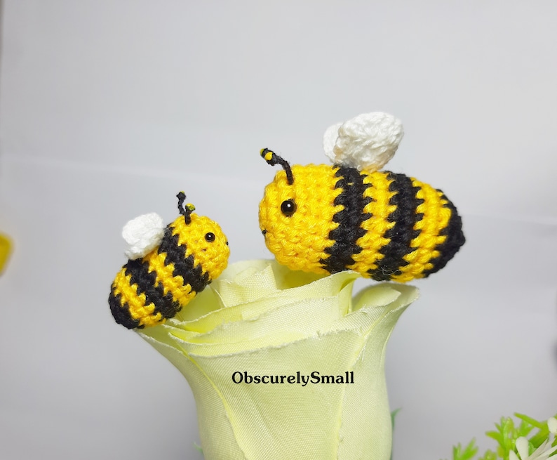 Op de afbeelding: Twee gehaakte hommels, een groter dan de andere, zitten op een witte roos. De bijen zijn geel en zwart met witte vleugels.