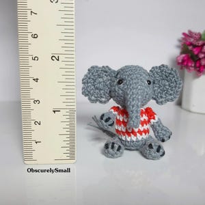 May include: A small gray crocheted elephant figurine wearing a red and white striped shirt. The elephant has black bead eyes and dark gray accents on its feet. A ruler is positioned next to the figure, showing it stands approximately 2 inches tall.