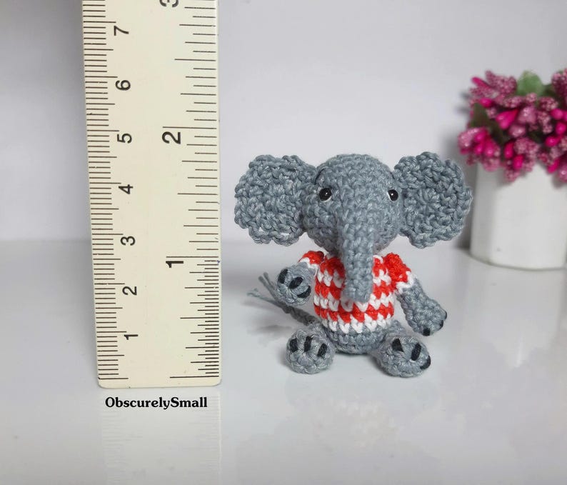 May include: A small gray crocheted elephant figurine wearing a red and white striped shirt. The elephant has black bead eyes and dark gray accents on its feet. A ruler is positioned next to the figure, showing it stands approximately 2 inches tall.
